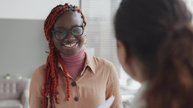 Back view of young African woman with colorful dreads and stylish eyeglasses standing in office and communicating with female colleague