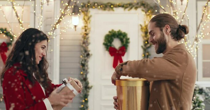 Close up portrait of cheerful Caucasian young married couple exchanging xmas presents while standing on decorated snowy street. Side view. Husband receives and opens gift box with surprised face.