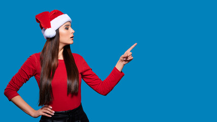 Fototapeta na wymiar Excited young woman in Christmas Santa hat showing or pointing with finger away on blue isolated background