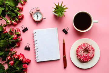 Feminine workplace with notepad, pen, alarm clock, flowers, cup of coffee and donut on pink background. Business, freelance and education concept. Morning concept. Top view, flat lay