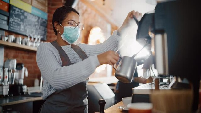 Beautiful Latin American Female Barista in Face Mask is Making a Cup of Cappuccino in Coffee Shop Bar. Social and Medical Health Restrictions Concept in a Loft-Style Cafe During Coronavirus Pandemic.