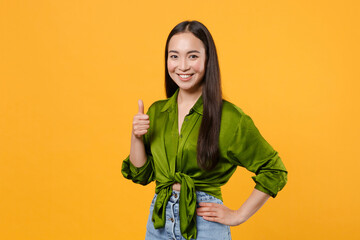 Smiling cheerful beautiful attractive young brunette asian woman 20s wearing basic green shirt standing showing thumb up looking camera isolated on bright yellow colour background, studio portrait.
