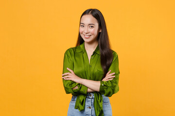 Smiling confident beautiful attractive young brunette asian woman wearing basic green shirt standing holding hands crossed looking camera isolated on bright yellow colour background, studio portrait.