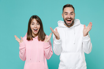 Excited surprised amazed young couple two friends man woman 20s wearing white pink casual hoodie keeping mouth open spreading hands isolated on blue turquoise colour wall background studio portrait.