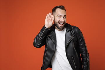 Curious excited young bearded man in basic white t-shirt black leather jacket standing try to hear you overhear listening intently looking aside isolated on orange colour background studio portrait.