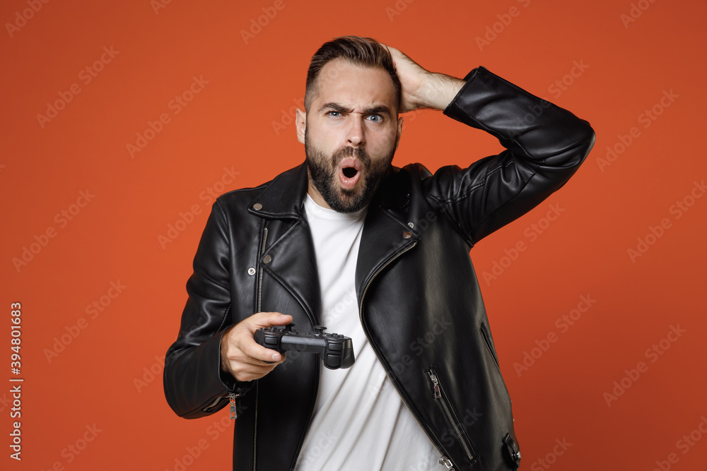 Wall mural Shocked worried young bearded man wearing casual basic white t-shirt, black leather jacket standing play pc game with joystick console put hand on head isolated on orange background studio portrait.