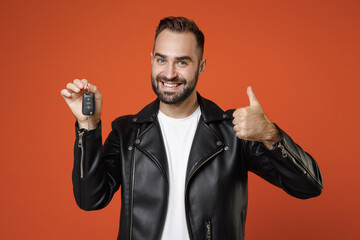 Smiling young bearded man 20s in basic white t-shirt, black leather jacket standing hold in hands car keys showing thumb up looking camera isolated on bright orange colour background studio portrait.
