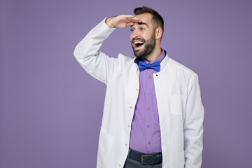 Excited young bearded doctor man in white medical gown holding hand at forehead looking far away distance isolated on violet background studio portrait. Healthcare personnel health medicine concept.