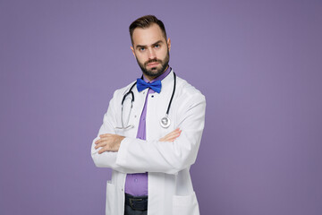 Dissatisfied young bearded doctor man in white medical gown looking camera holding hands crossed isolated on violet colour background studio portrait. Healthcare personnel health medicine concept.