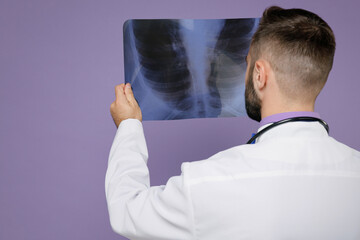 Back rear view of young bearded doctor man in medical gown stethoscope hold X-ray of lungs fluorography roentgen isolated on violet background studio. Healthcare personnel health medicine concept.