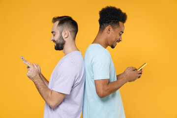 Side view of smiling young two friends european african american men 20s wearing violet blue casual t-shirts using mobile cell phone typing sms message isolated on yellow background studio portrait.
