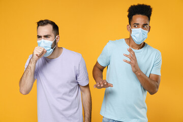 Sick worried young friends european african american men in purple blue t-shirts sterile face mask to safe from coronavirus coughing covering with hands isolated on yellow background studio portrait.