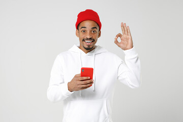 Cheerful young african american man 20s in basic casual streetwear hoodie standing using mobile cell phone typing sms message showing Ok gesture isolated on white colour background, studio portrait.