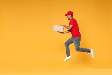 Full length delivery employee african man in red cap blank print t-shirt uniform work courier service on quarantine covid-19 virus concept jump hold cardboard box isolated on yellow background studio.