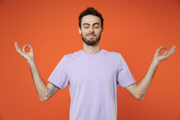 Fototapeta premium Smiling young bearded man 20s wearing casual violet t-shirt standing hold hands in yoga gesture, relaxing meditating, trying to calm down isolated on bright orange color background studio portrait.