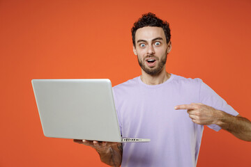 Shocked amazed young bearded man 20s in casual basic violet t-shirt standing working pointing index finger on laptop pc computer looking camera isolated on orange color background studio portrait.