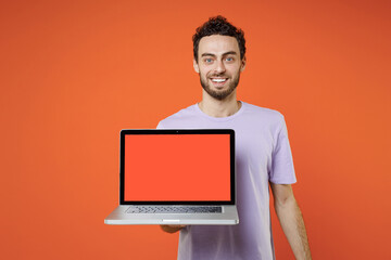 Smiling young bearded man 20s wearing casual basic violet t-shirt standing hold laptop pc computer with blank empty screen looking camera isolated on orange color wall background studio portrait.