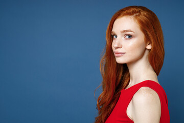 Side view of smiling charming beautiful pretty young redhead woman 20s wearing bright red elegant evening dress standing and looking camera isolated on blue color wall background studio portrait.
