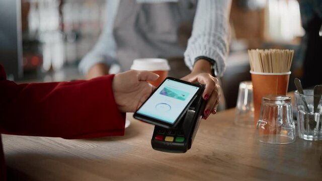 Close Up of a Feminine Hand Holding a Smartphone with an NFC Payment Technology Used for Paying for Take Away Coffee in a Cafe. Customer Uses Mobile to Pay for Latte Through a Credit Card Terminal.