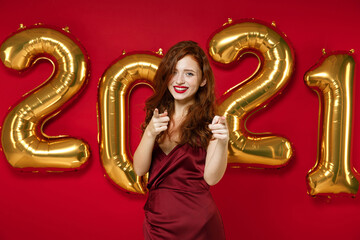 Smiling young redhead woman in elegant dress pointing index fingers on camera isolated on red background, golden numbers air balloons in studio. Happy New Year 2021 celebration holiday party concept.