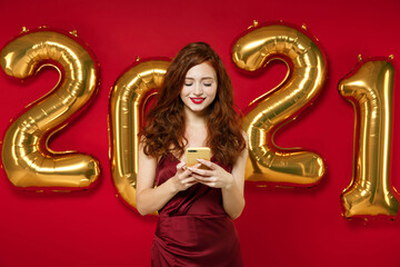 Smiling young redhead woman in elegant dress using mobile cell phone typing sms message isolated on red background, golden numbers air balloons. Happy New Year 2021 celebration holiday party concept.