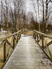 Frosty wooden bridge covered with white snowflakes over the frozen river and among tall birch trees in natural park of Pori, Finland, Europe. Cold winter snowy landscape, winterland.