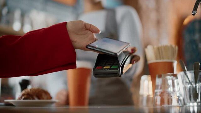 Close Up of a Feminine Hand Holding a Smartphone with an NFC Payment Technology Used for Paying for Take Away Coffee in a Cafe. Customer Uses Mobile to Pay for Latte Through a Credit Card Terminal.