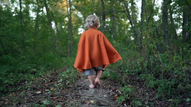 Little toddler boy cosplay gnome or hobbit in long cape walking barefoot in green forest. Halloween, kids concept. Amazing fairy tale character. Slow motion. 