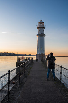 Man standing on a pier photographing a lighthouse at sunset.