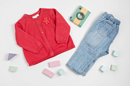 Composition with baby red cardigan, jeans, and toy photo camera on white background