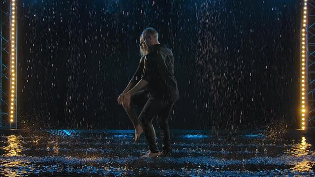 Active happy man and a woman dancing salsa together in a dark studio in the rain. A couple in black are enjoying the dance among rain drops and splashing water. Slow motion.
