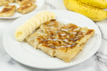 Banana paratha or pancake served in white plate with bananas in the background