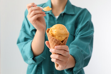 Woman eating yellow ice cream in wafer cone on light background, closeup