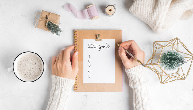 Desktop of woman hands with blank notepad writing 2021 goals text . Flat lay of white working table background with cup of coffee and Christmas decoration. Top view mock up 