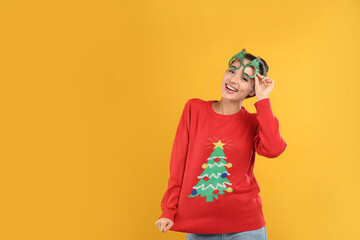 Young woman in Christmas sweater and party glasses on yellow background, space for text