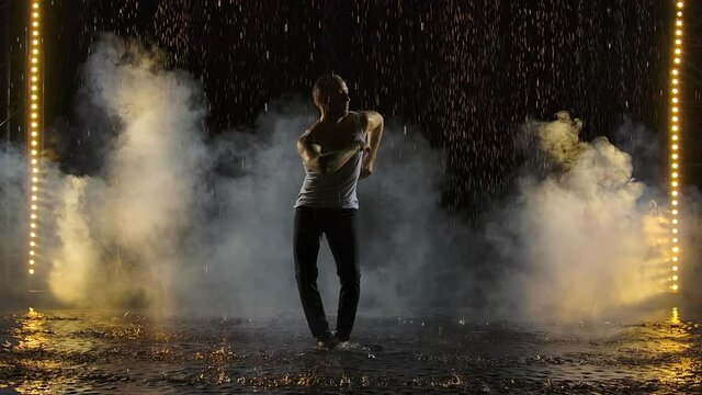 Silhouettes of male dancer dancing salsa elements on the water surface among rain drops and splashes of water. Solo performed by artist against the background of smoke and studio light. Slow motion