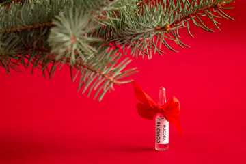 An ampoule of the COVID-19 coronavirus vaccine stands under a Christmas tree branch as a gift on a red background, copy space