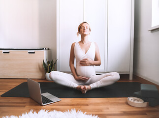 Pregnant woman practicing yoga online at home with laptop.