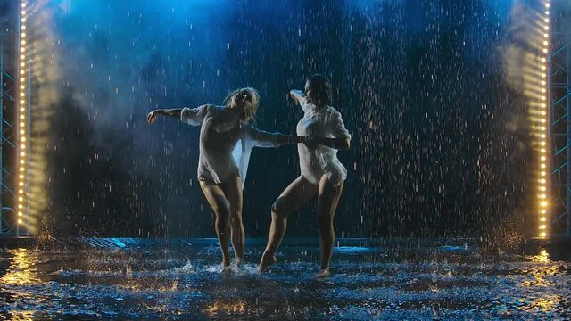 Two young sexy women passionately dancing salsa elements in raindrops and smoke. Women's silhouettes in wet bodysuits and white shirts move erotically in slow motion.