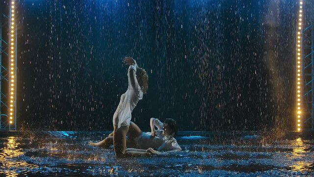 Sexy beautiful women erotically move on the surface of the water creating a lot of splashes. Two females in wet clothes under streams of rain on a smoky studio background. Slow motion.