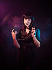 girl on a black background with pink-blue light and a slight haze. She is holding a telephone set, the receiver of which is at her ear.