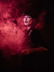 a girl in a harness in red light and smoke looks at the camera and holds a phone receiver in her...