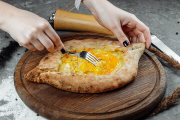 close-up of female hands with a knife and a fork mix the egg cheese in Adjarian khachapuri on a plate in a restaurant. Georgian national food