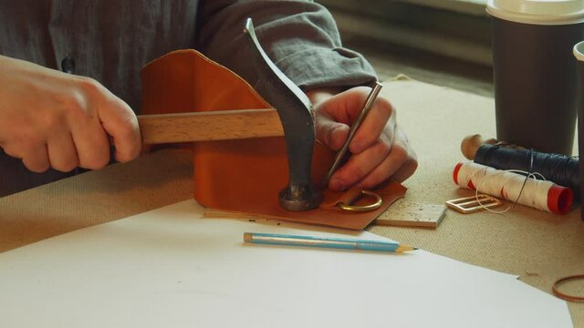 With a core and hammer, the tanner punches holes for sewing an exclusive bag made of genuine leather. Slow motion.