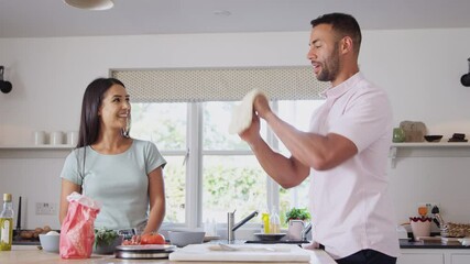 Couple at home in kitchen preparing homemade pizza as man spins base in the air - shot in slow motion