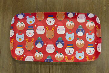 A red rectangular plate on wooden background. Plate has holiday season, christmas and new year related figures on it. This include, snow man, penguin, santa claus, bear, rein deer with antlers.