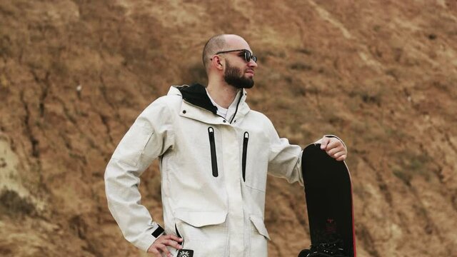 Handsome bald bearded man with sunglasses posing with sandboard in desert or sand quarry