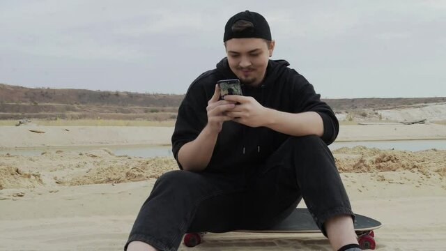 A Young Man sits on a Skateboard and uses phone in desert