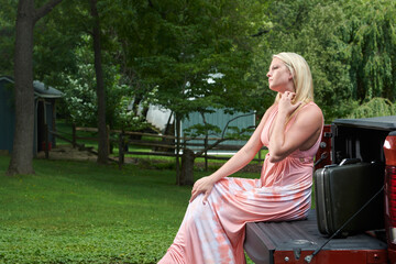 Sexy young blonde woman poses on or near farm, sitting on truck tailgate, wearing long flowing pink...