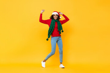 Fototapeta na wymiar Fun portrait of happy African American woman in Christmas attire smiling and raising hands on isolated yellow studio background